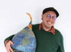 Stace holds a large papier mâché chicken under one arm like a football. He is laughing at the camera.